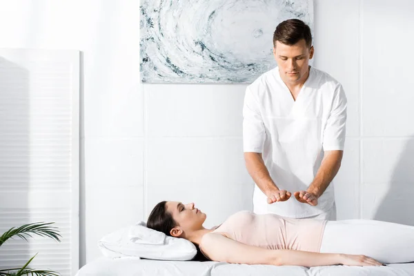 Focused healer standing near woman on massage table and holding hands above her stomach — Stock Photo