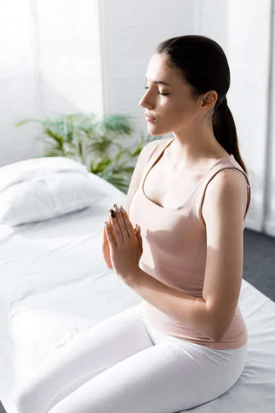 Femme assise sur une table de massage les yeux fermés et montrant un signe namaste à la clinique — Photo de stock