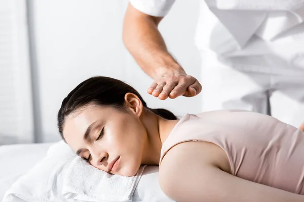 Vista recortada del sanador de pie cerca de la mujer acostada con los ojos cerrados en la mesa de masaje y cogida de las manos por encima de su cuerpo - foto de stock