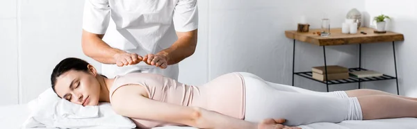 Plano panorámico del sanador de pie cerca de la mujer acostada con los ojos cerrados en la mesa de masaje y cogida de la mano por encima de su cuerpo - foto de stock