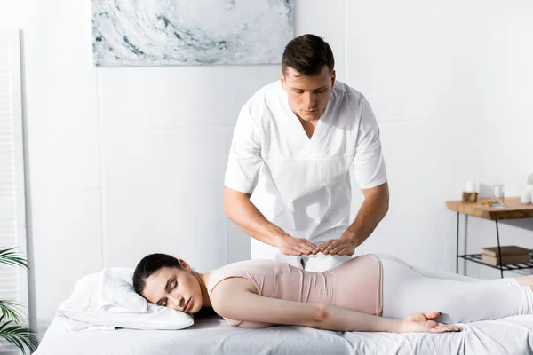 Sanadora enfocada de pie cerca de una mujer acostada sobre una mesa de masaje con los ojos cerrados y sosteniendo las manos sobre su cuerpo - foto de stock