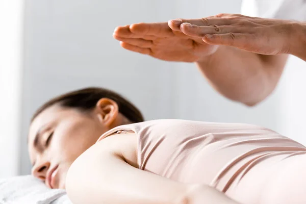 Vista recortada del sanador de pie cerca de la mujer acostada con los ojos cerrados y sosteniendo las manos sobre su cuerpo - foto de stock