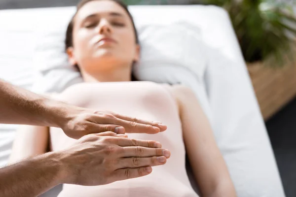 Vista ritagliata del guaritore in piedi vicino alla donna sdraiata con gli occhi chiusi sul tavolo da massaggio e tenendo le mani sopra il suo corpo — Foto stock