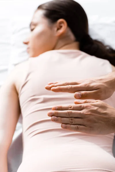 Vista recortada del sanador de pie cerca de la mujer acostada con los ojos cerrados y sosteniendo las manos sobre su cuerpo - foto de stock