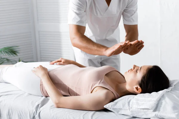 Vista recortada del sanador de pie cerca de la mujer en la mesa de masaje y la limpieza de su aura - foto de stock