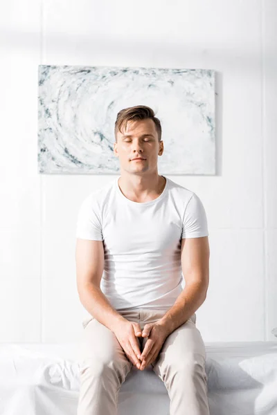 Vorderansicht eines Mannes in weißem T-Shirt, der mit geschlossenen Augen auf einem Massagetisch in der Klinik sitzt — Stockfoto