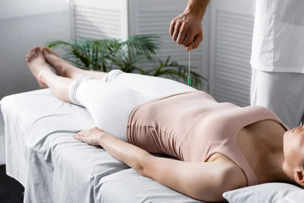 Cropped view of hypnotist standing near woman on massage table and holding green stone — Stock Photo