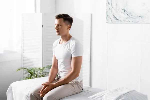 Hombre en camiseta blanca sentado en la mesa de masaje con los ojos cerrados en la clínica - foto de stock