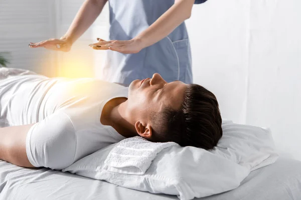 Vista parcial del sanador de pie cerca del hombre en la mesa de masaje y sosteniendo las manos por encima de su cuerpo - foto de stock