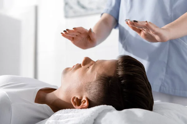 Partial view of healer standing near man on pillow and holding hands above his body — Stock Photo