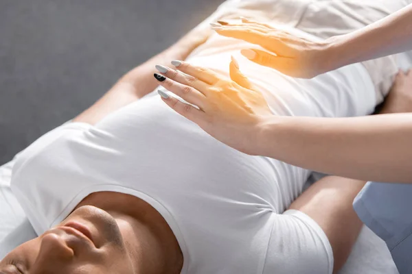 Visão parcial do curandeiro em pé perto do homem na mesa de massagem e segurando as mãos acima de seu corpo — Fotografia de Stock