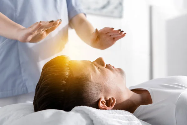 Partial view of healer standing near man on pillow and holding hands above his body — Stock Photo