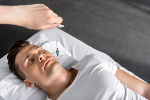 Vista cortada de hipnotizador em pé perto do homem na mesa de massagem e segurando pedra verde — Fotografia de Stock