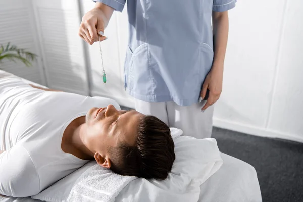 Cropped view of hypnotist standing near man on massage table and holding green stone — Stock Photo