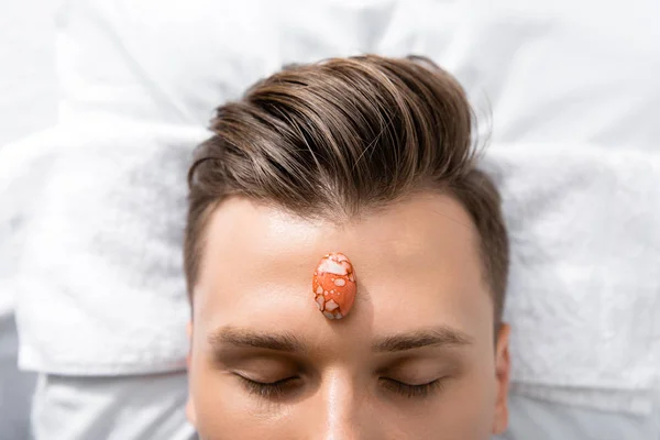 Vista recortada del hombre acostado en la almohada con los ojos cerrados con piedra en la frente - foto de stock