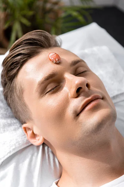 Smiling young man lying on pillow with closed eyes with stone on forehead — Stock Photo