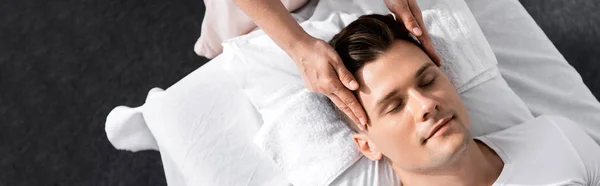 Panoramic shot of masseur standing near man with closed eyes and touching his face — Stock Photo