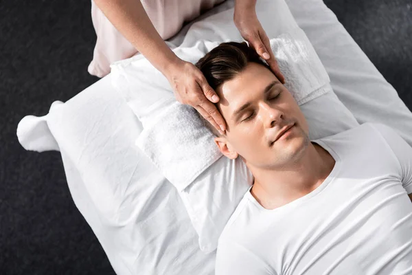Обрезанный вид массажера, стоящего рядом с человеком с закрытыми глазами и трогающего его лицо — стоковое фото