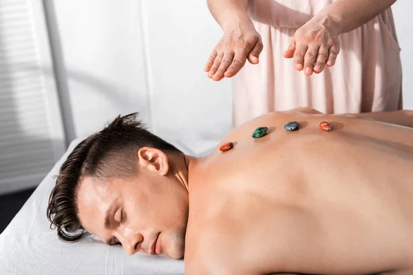 Vista recortada del sanador de pie cerca del hombre sin camisa acostado con los ojos cerrados con piedras en la espalda - foto de stock