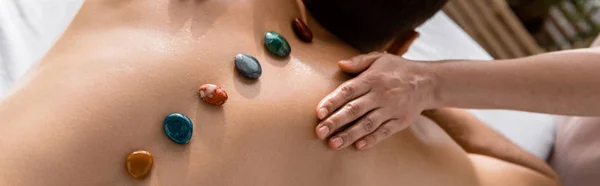 Panoramic shot of masseur and shirtless man with stones on back — Stock Photo