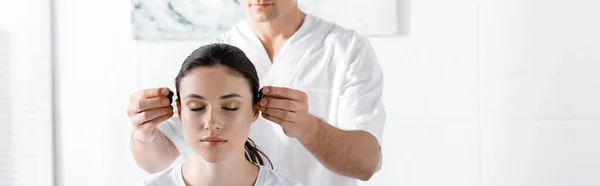 Panoramic shot of woman sitting with closed eyes while healer holding stones near her ears — Stock Photo