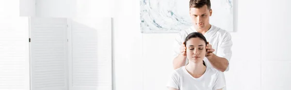 Panoramic shot of young woman sitting with closed eyes while masseur touching her ears — Stock Photo