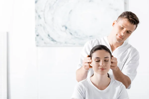 Jeune femme assise avec les yeux fermés tandis que masseur toucher ses oreilles — Photo de stock