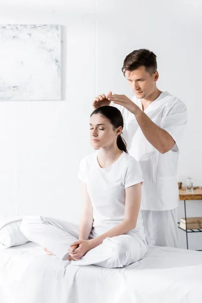 Woman sitting on massage table with closed eyes while healer holding hands above her head — Stock Photo