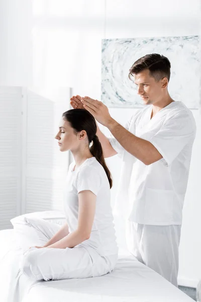 Woman sitting on massage table with closed eyes while healer holding hands above her head — Stock Photo