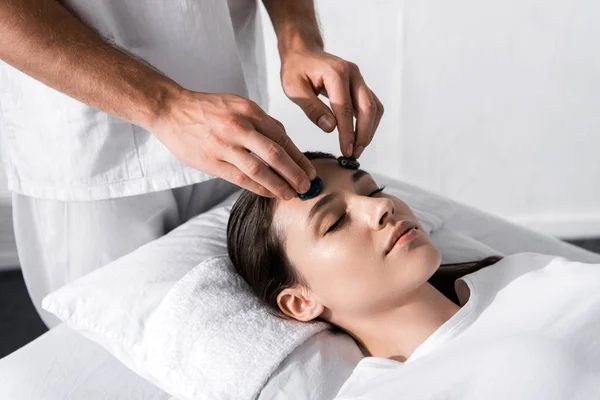 Vista recortada de curandero de pie cerca de la mujer con los ojos cerrados y utilizando piedras - foto de stock