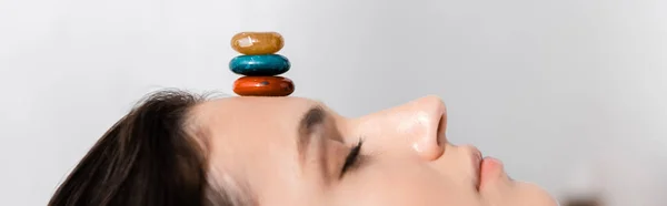 Panoramic shot of woman lying with closed eyes with colorful stones on forehead on grey — Stock Photo