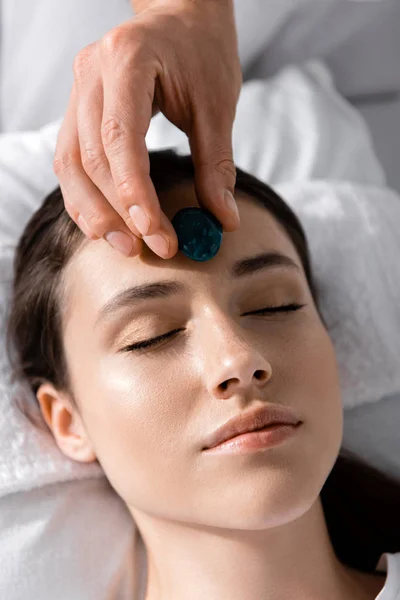 Cropped view of healer standing near woman lying on pillow with closed eyes and using stone — Stock Photo