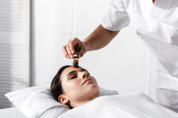 Vista recortada de curandero de pie cerca de la mujer acostada en la almohada con los ojos cerrados y utilizando piedras - foto de stock