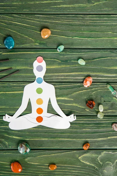 Vista superior de la figura de papel en forma de persona con chakras en pose de loto, palos de aroma y piedras de colores en la superficie de madera - foto de stock