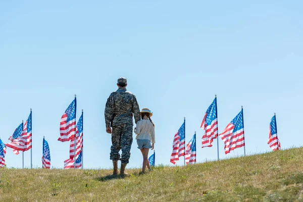Vista trasera del padre en uniforme militar cogido de la mano con el niño cerca de banderas americanas - foto de stock