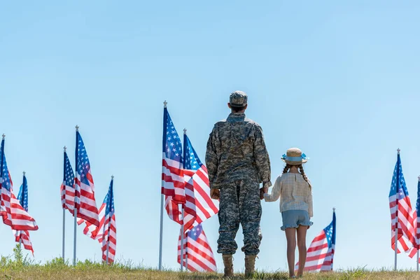 Vue arrière du père en uniforme militaire tenant la main avec un enfant près des drapeaux américains — Photo de stock