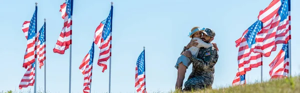 Panoramic shot of kid hugging father in military uniform near american flags — Stock Photo