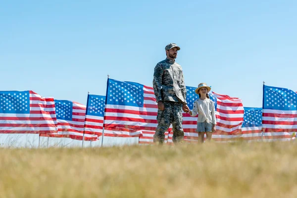Messa a fuoco selettiva di papà in uniforme militare che si tiene per mano con bambino vicino alle bandiere americane — Foto stock