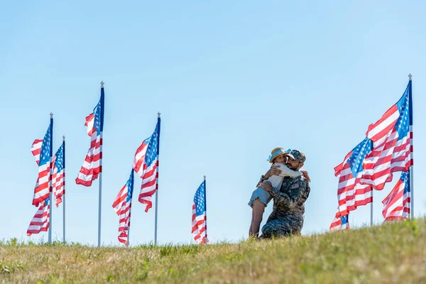 Garoto bonito abraçando o pai em uniforme militar perto de bandeiras americanas — Fotografia de Stock