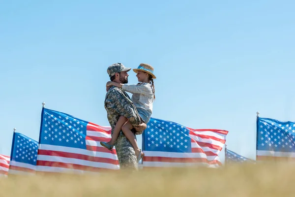 Вибірковий фокус військового батька, який тримає в руках веселу дитину біля американських прапорів — стокове фото