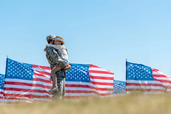Вибірковий фокус військового чоловіка в уніформі, що тримається в руках весела дитина біля американських прапорів — стокове фото
