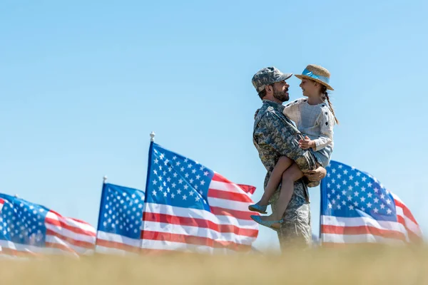 Вибірковий фокус людини у військовій формі, що тримається в руках весела дитина біля американських прапорів — стокове фото