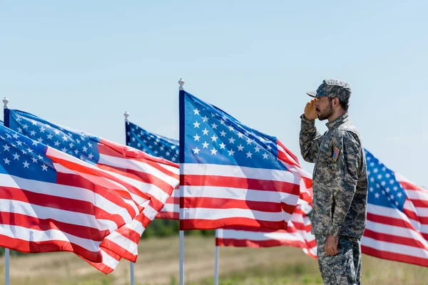 Hombre en uniforme militar y gorra de pie y dando saludo cerca de banderas americanas — Stock Photo