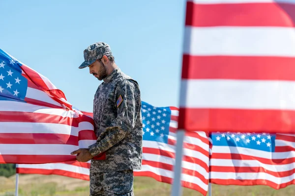 Selective focus of man in military uniform and cap standing and touching american flag — Stock Photo