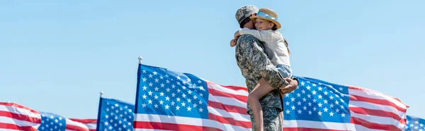 Panoramic shot of military man holding in arms cheerful child near american flags — Stock Photo