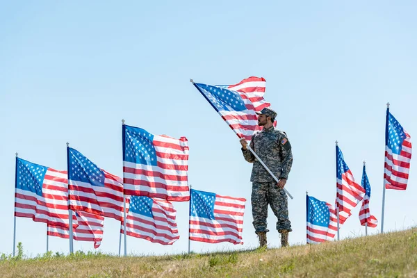 Foco seletivo do homem bonito em uniforme militar e tampa de pé e segurando bandeira americana — Fotografia de Stock