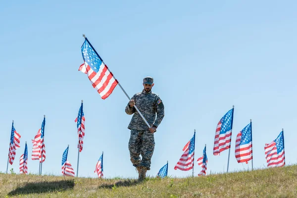 Guapo soldado en uniforme militar y gorra sosteniendo bandera americana mientras está de pie en la hierba - foto de stock