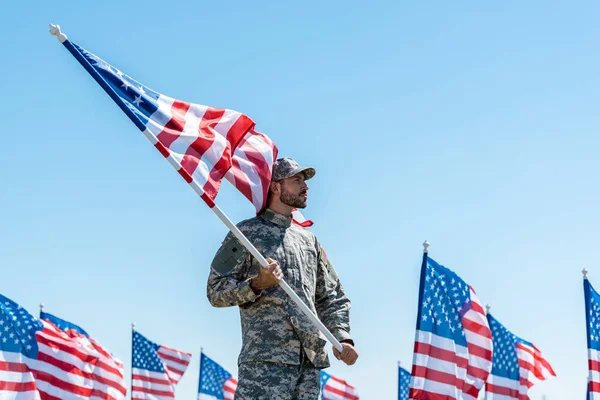 Bel homme en uniforme militaire et casquette tenant le drapeau américain tout en se tenant contre le ciel bleu — Photo de stock