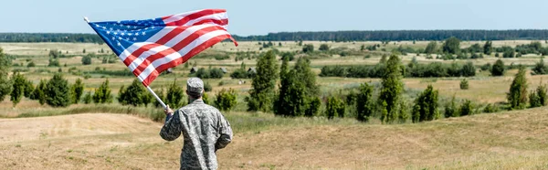 Panoramic shot of military man standing near trees and holding american flag — Stock Photo