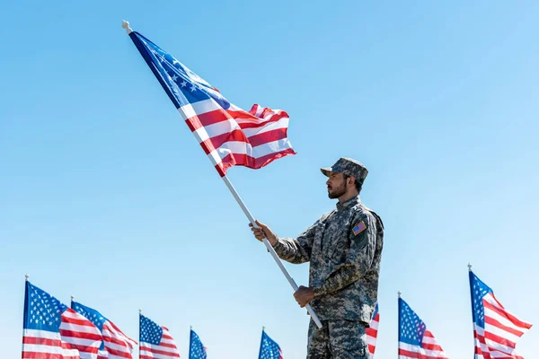 Handsome military man in uniform and cap holding american flag while standing against blue sky — Stock Photo